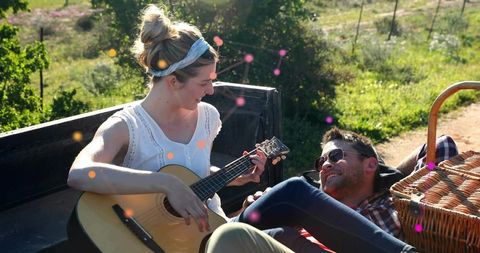 Couple playing guitar in truck bed relaxing in sunlit meadow with picnic basket