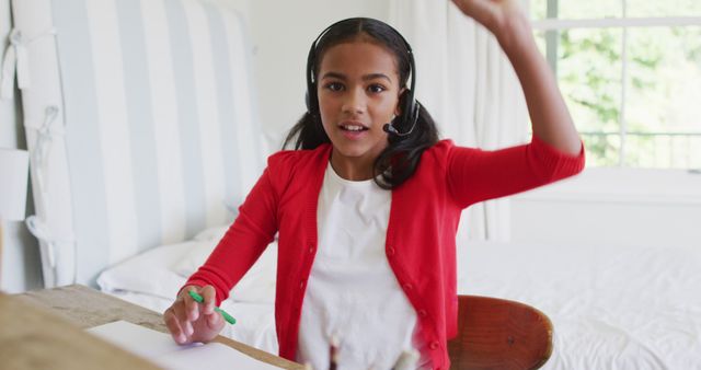 Black girl attending virtual class at home, actively participating with raised hand and wearing headphones. Ideal for use in education blogs, online learning platforms, and remote schooling advertisements to depict engaged students in a home learning environment.