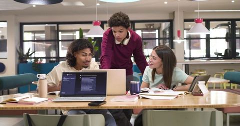 Three students collaborating around laptops in modern campus study lounge for teamwork