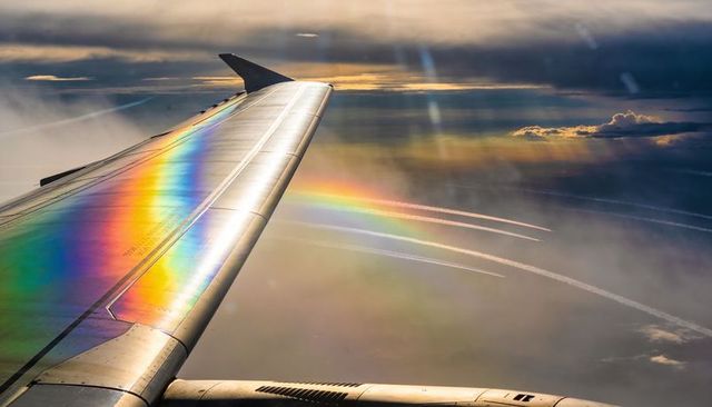 Iridescent airplane wing casting rainbow bands over clouds and contrails at golden hour