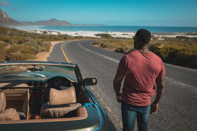 African American man standing next to a convertible car on a winding coastal highway. The scene captures the essence of a summer road trip with scenic views of the ocean and surrounding nature. Ideal for use in travel blogs, vacation advertisements, lifestyle magazines, and automotive promotions.