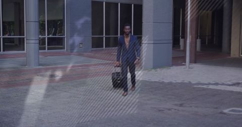 Urban business traveler walking across glass-front plaza pulling wheeled suitcase