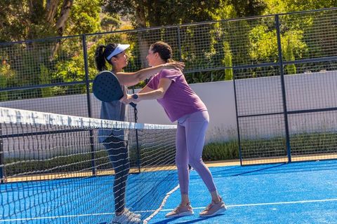 Female players greeting across net on blue paddle court during sunny outdoor match