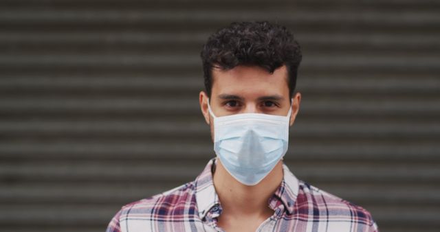 Young man with curly hair standing wearing face mask for health safety. Ideal for content about covid-19 prevention, public health measures, and pandemic lifestyle. Captures concept of personal safety, health awareness, and urban living during a pandemic.