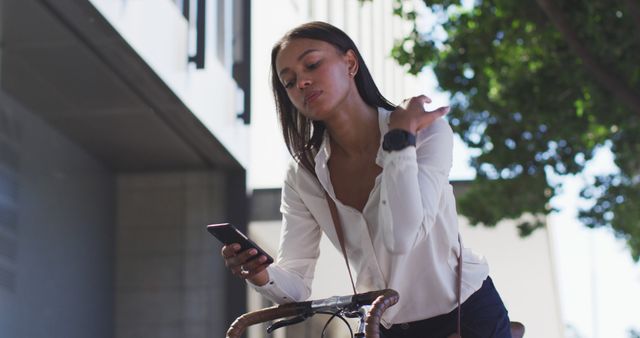 Businesswoman checks phone while riding bicycle in an urban area. Ideal for themes of modern communication, sustainable commuting, professional lifestyle, and urban living.