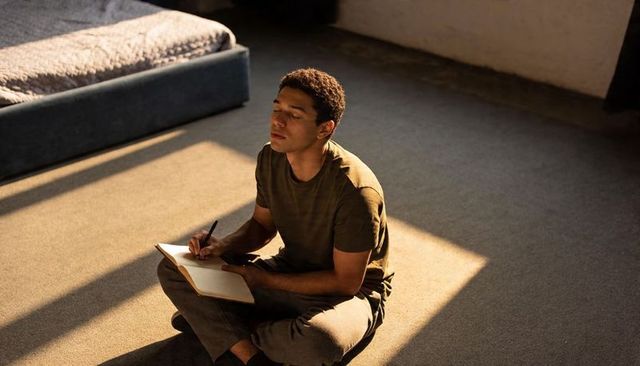 Adult man journaling on bedroom floor in warm sun patch, contemplative minimalist moment