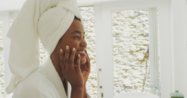 Reflecting moment of a woman during her skincare routine in a modern, bright bathroom. Wearing bathrobe and towel turban, gently touching her face. Perfect for ads focused on beauty products, self-care routines, or relaxed living styles.