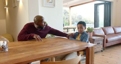 African american father reassuring upset son at wooden dining table in cozy open-plan home