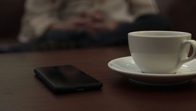 Porcelain cup and saucer resting on wooden table with smartphone and blurred person in lounge
