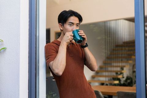 Asian man sipping coffee in doorway wearing rust polo and smartwatch, modern home interior
