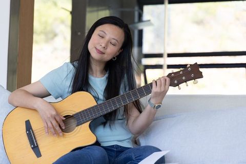 Relaxed asian woman sitting on sofa playing acoustic guitar while reading music sheet