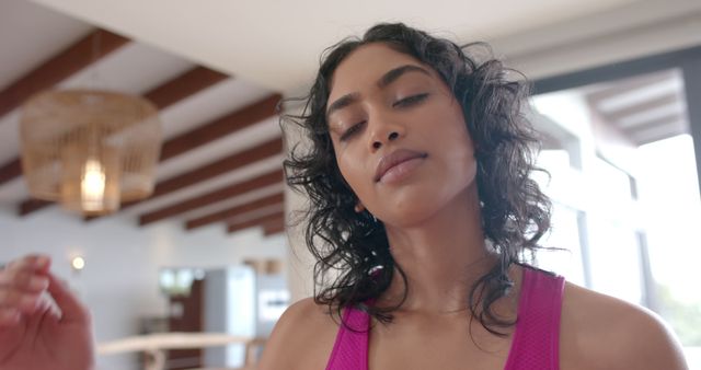 Young woman in pink athletic wear looks relaxed and satisfied after a workout session in a stylish, well-lit home interior. Her eyes are closed, lips slightly curved into a smile, conveying sense of achievement and tranquil energy. Ideal for themes related to fitness, health, relaxation, wellness retreats, and modern lifestyle.