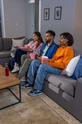 Diverse friends sitting on sectional sofa watching movie night holding popcorn and drinks