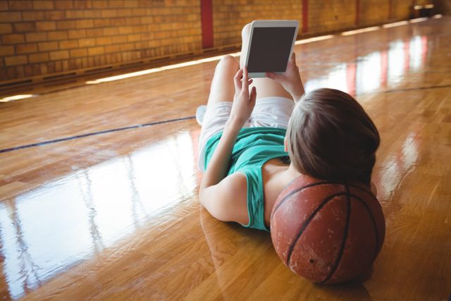 Woman lying on basketball court using digital tablet, resting head on basketball. Ideal for themes of sports and technology, leisure activities, fitness lifestyle, and modern relaxation. Suitable for articles, blogs, and advertisements related to sports, technology, and casual lifestyle.