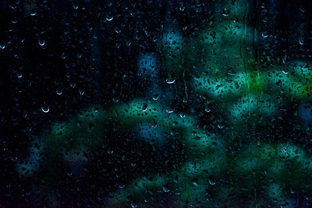 Raindrops cover a darkened window pane with blurred green lights visible in the background. Ideal for use in themes involving rain, melancholy moods, or nature's abstract beauty. This can also be used in weather-related articles or as background art for blogs and websites.