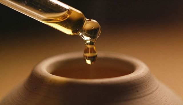 Golden oil dropper releasing single droplet into ceramic bowl under warm light
