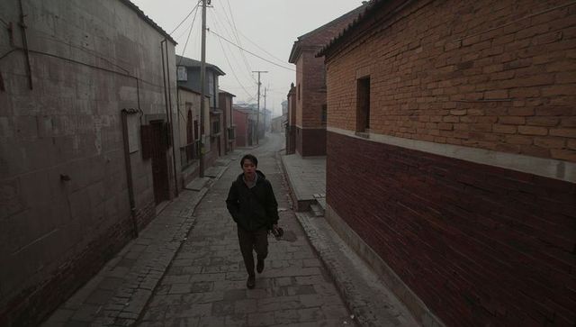 Solitary man walking uphill on foggy cobblestone street between brick houses holding shoes