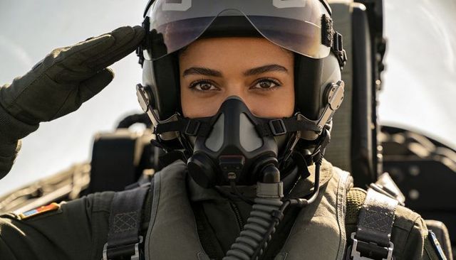 Female military pilot saluting in jet cockpit wearing helmet and oxygen mask