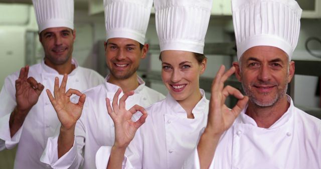 Four chefs standing in kitchen wearing white chef uniforms and hats, smiling and making OK gesture with hands. Perfect for illustrating professional teamwork in culinary arts, restaurant management content, or advertisements promoting food services and hospitality.