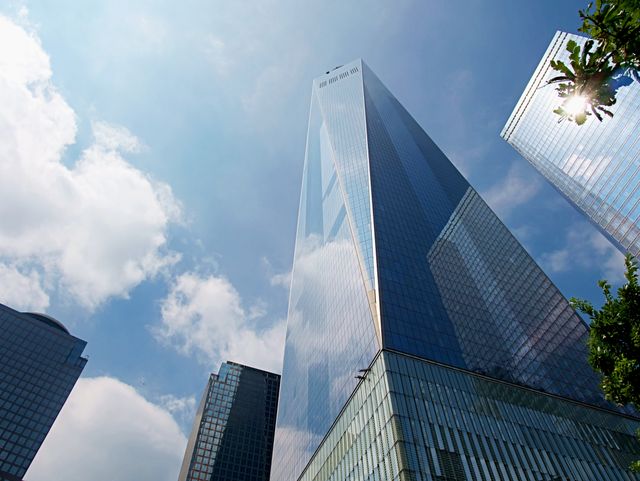 One World Trade Center towering against a backdrop of blue sky with clouds. The glass and steel structure reflects surrounding buildings, creating a modern urban scene. Ideal for use in content related to architecture, business districts, urban lifestyle, travel, and tourism in New York City.