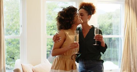 Same-Sex Couple Kissing Cheek While Holding Champagne Flutes Celebrating at Home Together