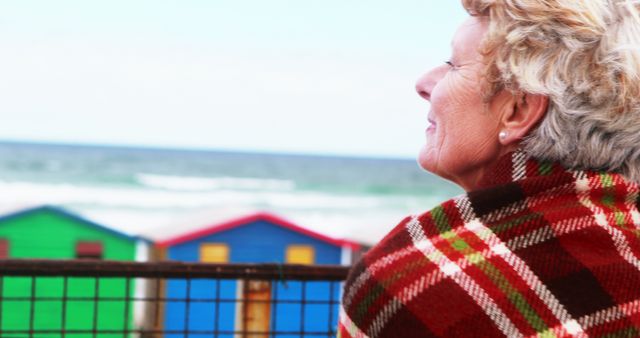 A senior Caucasian woman wrapped in a plaid blanket gazes out at colorful beach huts and the ocean, with copy space. Her contemplative expression suggests a moment of peaceful reflection or nostalgia by the seaside.