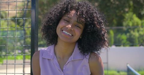 Curly-haired woman smiling by metal fence wearing lavender sleeveless blouse, relaxed portrait