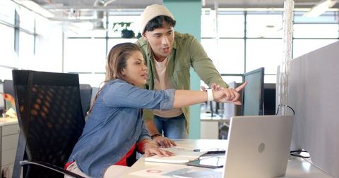 Diverse coworkers collaborating at modern open office desk pointing at laptop screen