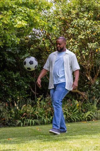 African american man playing soccer in backyard garden kicking ball on green lawn