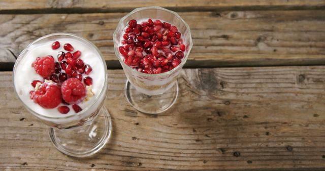 Two glasses of yogurt parfait with fresh raspberries and pomegranate seeds are presented on a rustic wooden table, with copy space. The vibrant red berries add a pop of color and suggest a healthy and refreshing snack option.