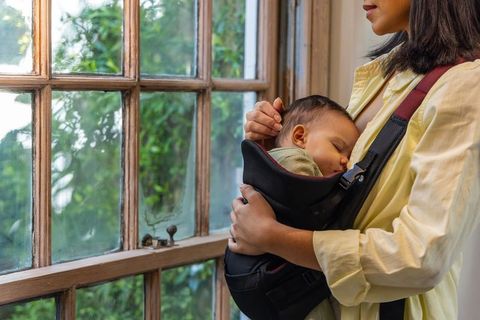 Mother cradling sleeping baby in front baby carrier by sunlit wooden window