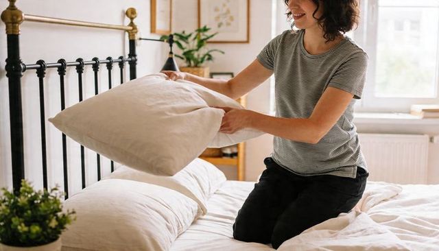 Woman fluffing pillow on made bed in bright minimalist bedroom with iron headboard
