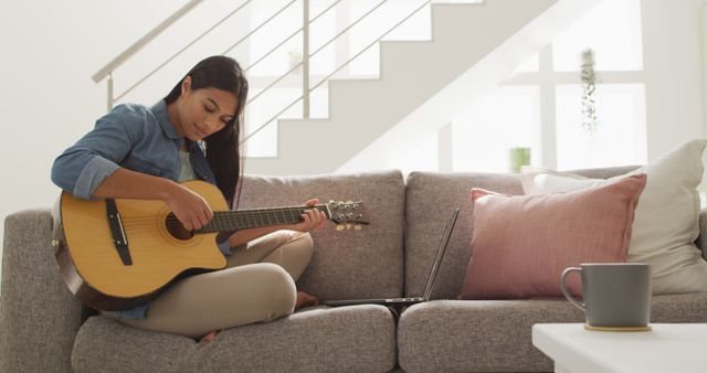Young woman sitting on a cozy gray couch in a modern living room playing an acoustic guitar. Her laptop is open on the couch beside her. The setting is relaxed, with soft lighting highlighting the interior. Perfect for use in themes related to home leisure, music practice, work-from-home lifestyles, creativity, and wellbeing.