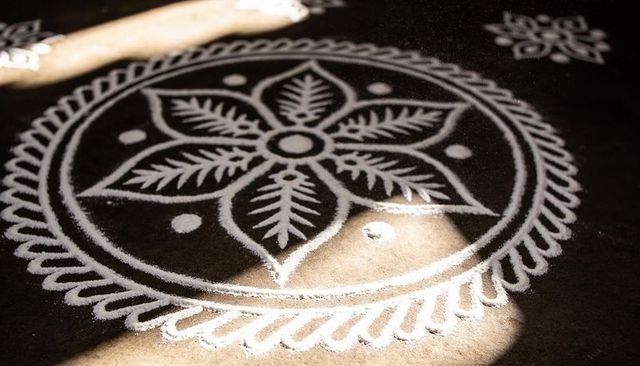 White floral rangoli mandala on dark veranda floor with sunlight patches and wooden stick