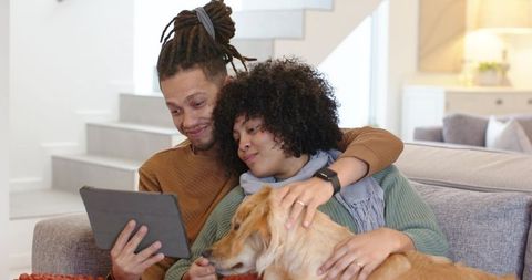 Couple cuddling with golden retriever while browsing tablet on cozy living room sofa