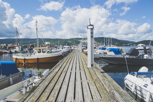 Wooden dock stretching into a marina filled with various boats. Calm water and sailboats create a scenic view, with green hills in background. Ideal for use in travel brochures, websites about outdoors or boating, or blogs on scenic destinations.