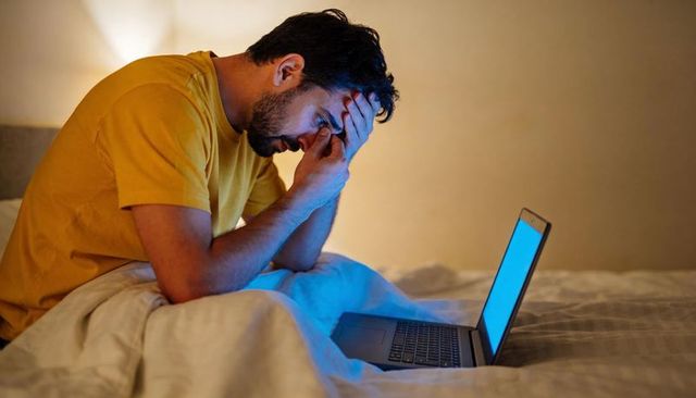 Man rubbing eyes late night on bed while staring at laptop blue screen glow