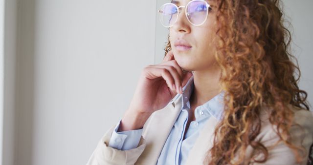 This image of a young female professional with curly hair and glasses can be used to represent confident businesswomen, female leadership, or entrepreneurial spirit. Suitable for corporate websites, business blogs, and professional magazines emphasizing women in the workplace and thoughtful leadership.