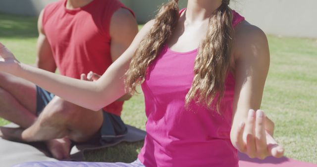 Man and woman are sitting on yoga mats practicing yoga meditation in park. Woman in foreground is wearing pink activewear while airing long hair. Man is slightly behind her wearing red tank top. Suitable for use in fitness and wellness articles, marketing materials for yoga classes, outdoor exercise promotions, mental health awareness campaigns.