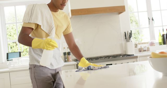 Young man wearing yellow gloves cleaning kitchen countertop. This image signifies cleanliness and domestic chores, showcasing a person taking care of their home environment. Useful for articles on home cleanliness, pest control, lifestyle blogs, domestic tips, or advertisements for cleaning supplies.