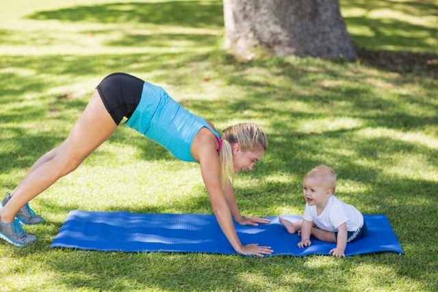 Mother doing a yoga workout on a blue mat in a park, bonding with her baby nearby. Perfect for promoting family health and fitness, outdoor activities, parenting tips, and active lifestyles.
