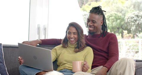 Multicultural couple relaxing on sofa using laptop and sipping coffee in sunlit living room