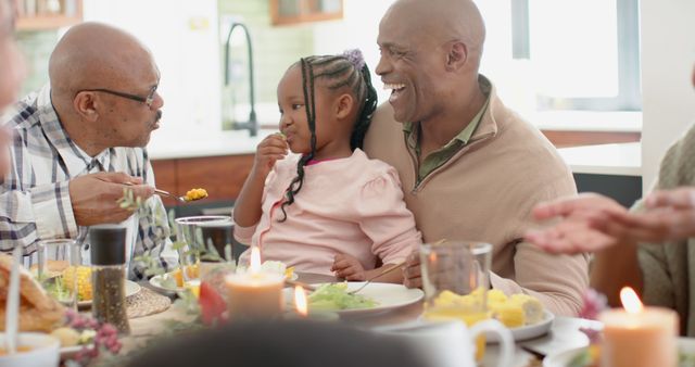 Three generations of an African American family are gathered around the dinner table, celebrating Thanksgiving. A young girl smiles while being fed by her grandfather, with her father joining in the cheerful moment. Use this to depict family bonding, holiday celebrations, warmth of home, or generational love.