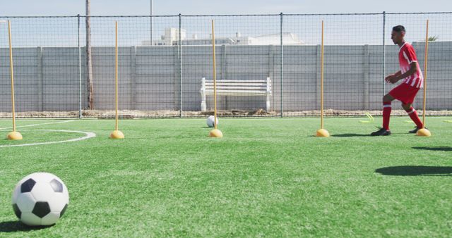 A football player in red jersey practicing training drill on a grass field with cones set up. Ideal for illustrating soccer training, sports academies, athletic practice sessions, and fitness routines.