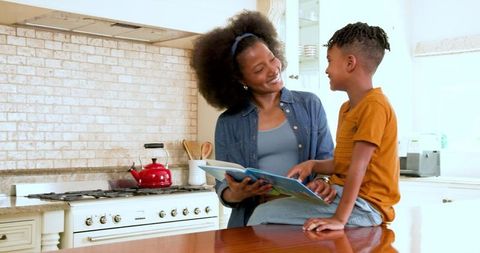 Happy Mother Reading Book with Her Son at Home Dining Table