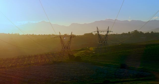 Sunset illuminating rural landscape with power lines stretching across fields towards distant mountains. Showcases harmony between nature and infrastructure. Ideal for topics on renewable energy, rural living, ecological balance, and sustainable development ideas.