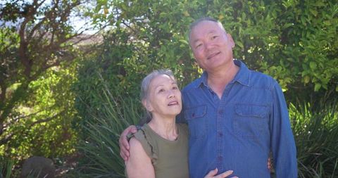 Senior asian couple standing in sunlit garden looking up together embracing warm moment