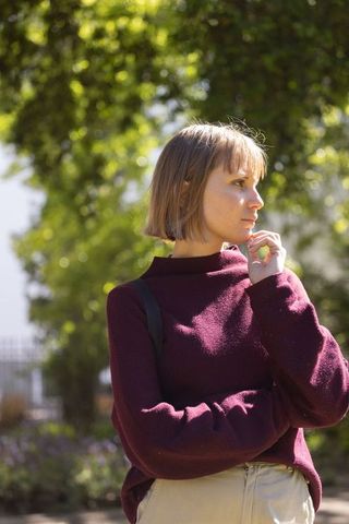 Young woman standing under dappled sunlight in garden wearing maroon sweater and backpack