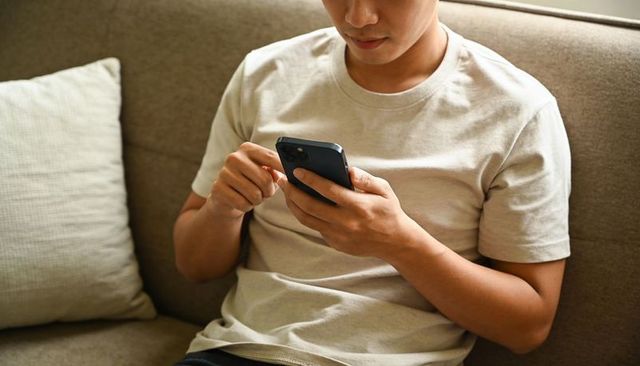 Young man scrolling smartphone on cozy sofa, checking messages and notifications at home