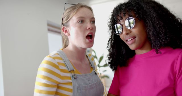 Two young women, with one wearing a striped shirt and overalls, and the other in a pink shirt and sunglasses, socialize indoors. Ideal for themes of friendship, casual lifestyle, social interaction, and youth engagement.